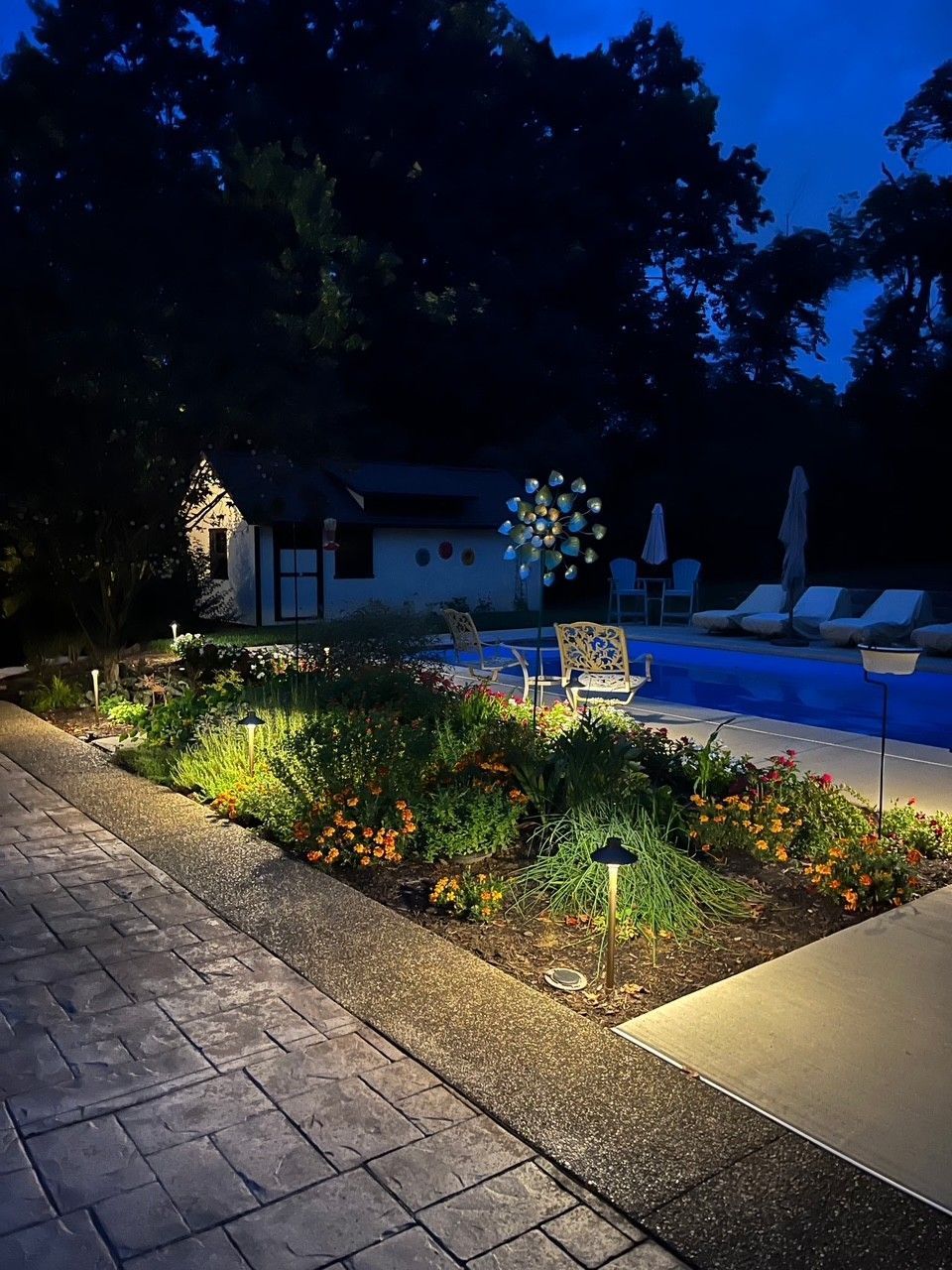 Nighttime patio garden with lit flower beds, stone walkway, and swimming pool lounge chairs