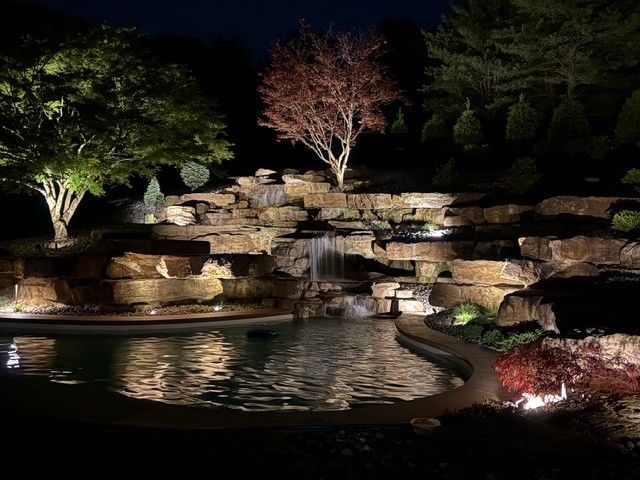 Illuminated rock waterfall and pond at night, with trees and landscaped greenery.