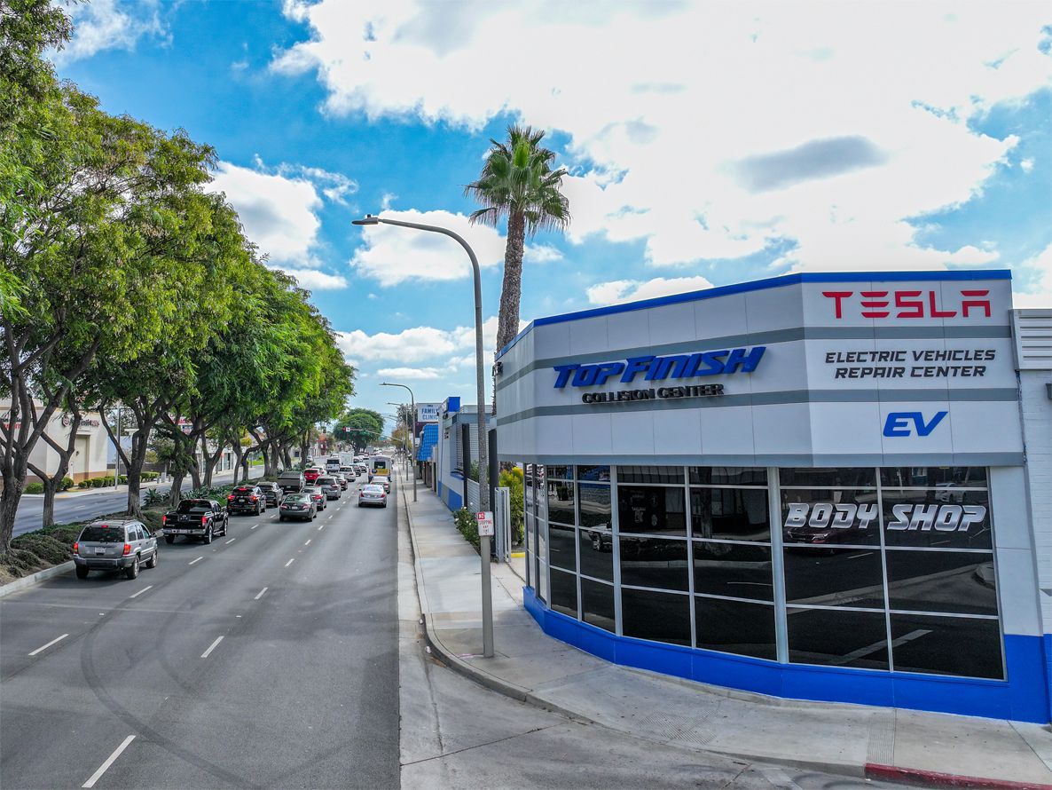 Tesla electric vehicle repair center; cars on street, palm tree, blue and white building.