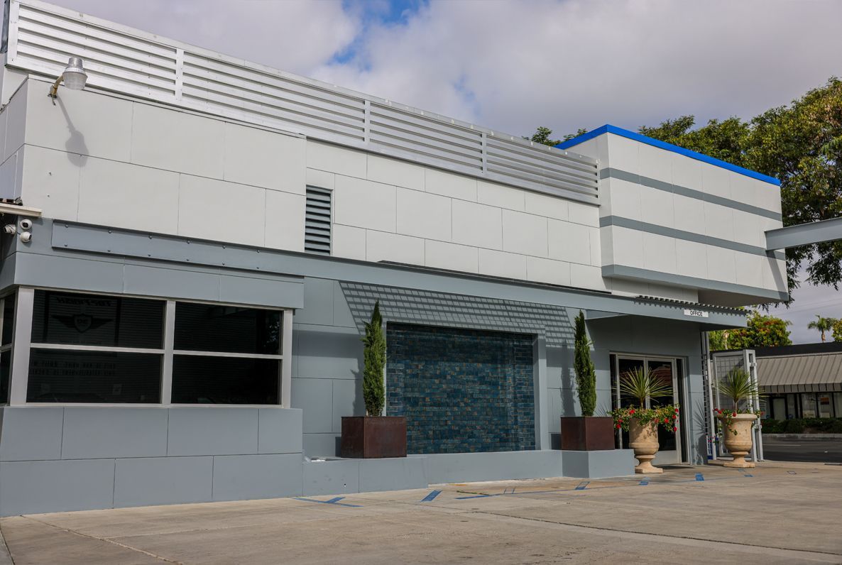 Gray and white modern building with tinted windows, potted plants, and a blue sky background.