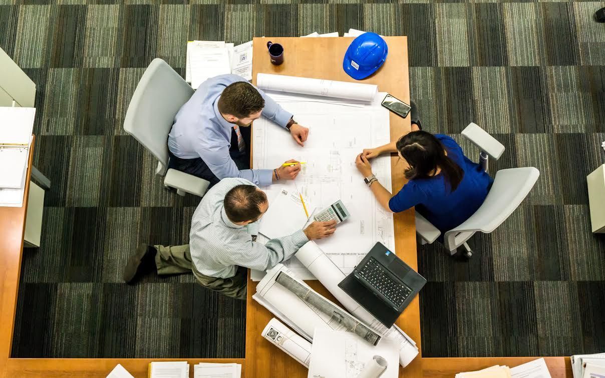 Three people collaborate over blueprints at a desk with a blue hard hat.