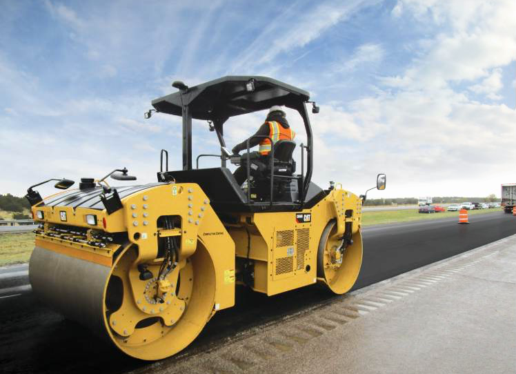 Yellow road roller compacting asphalt on a highway. A worker is in the cab.