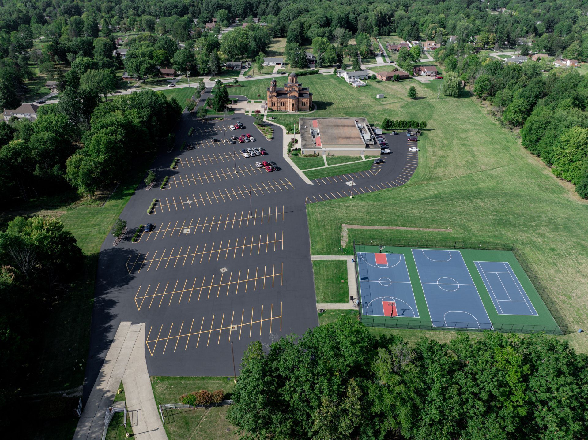 Aerial view of a parking lot, playground, and multi-sport courts next to a building and trees.