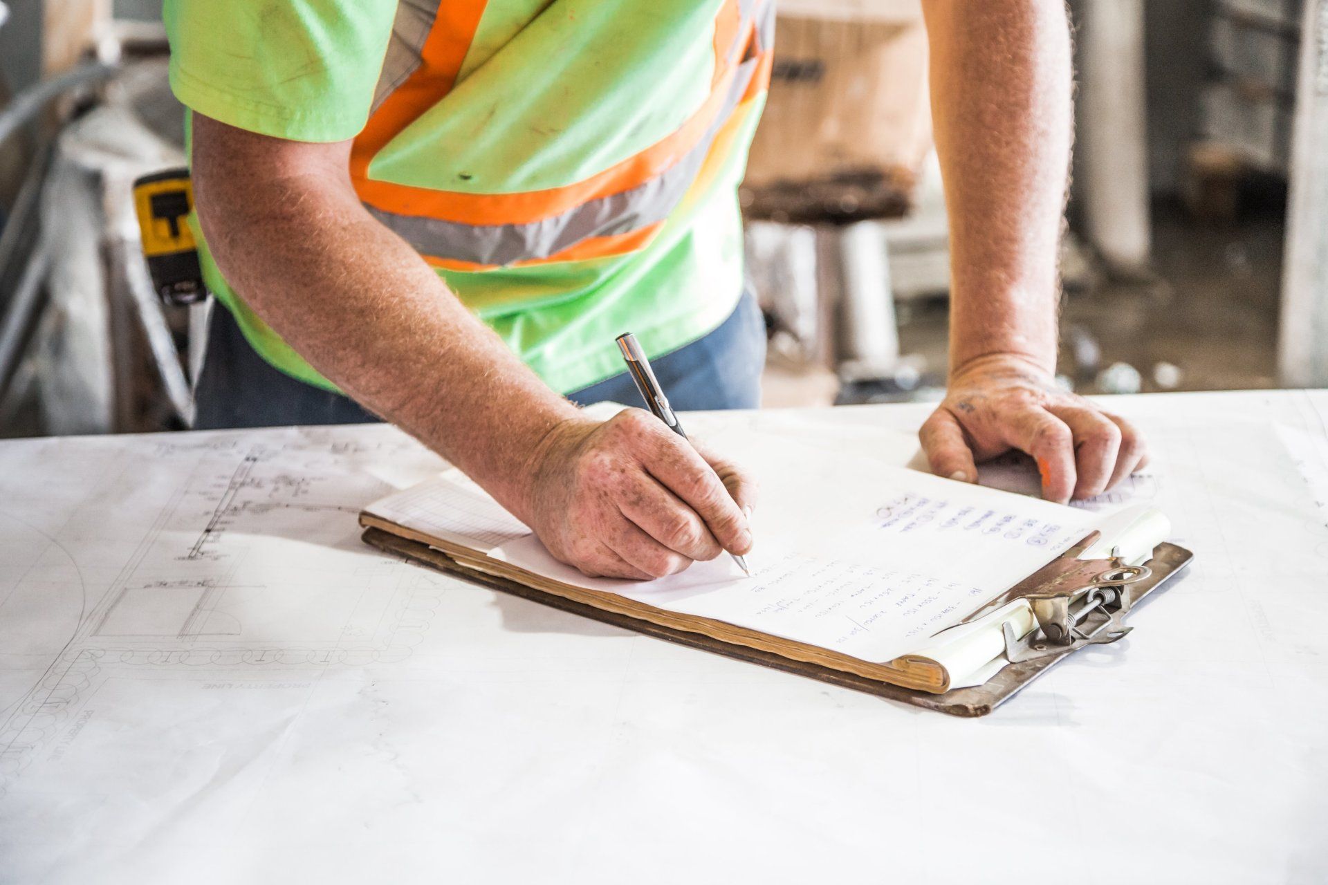 Construction worker in a safety vest writing on a clipboard, standing at a table with blueprints.