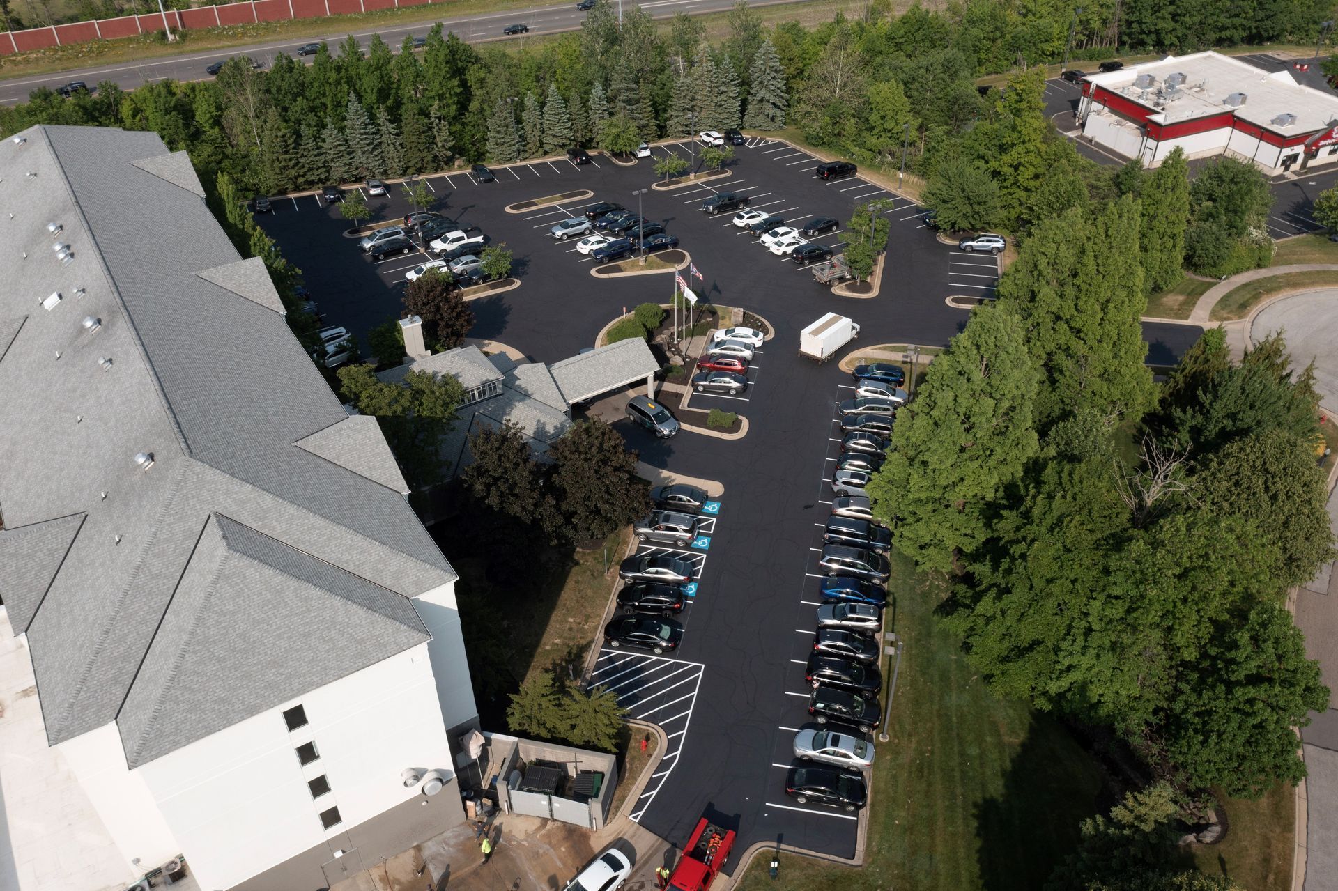 Aerial view of a parking lot filled with cars. A building is on the left, trees on the right.