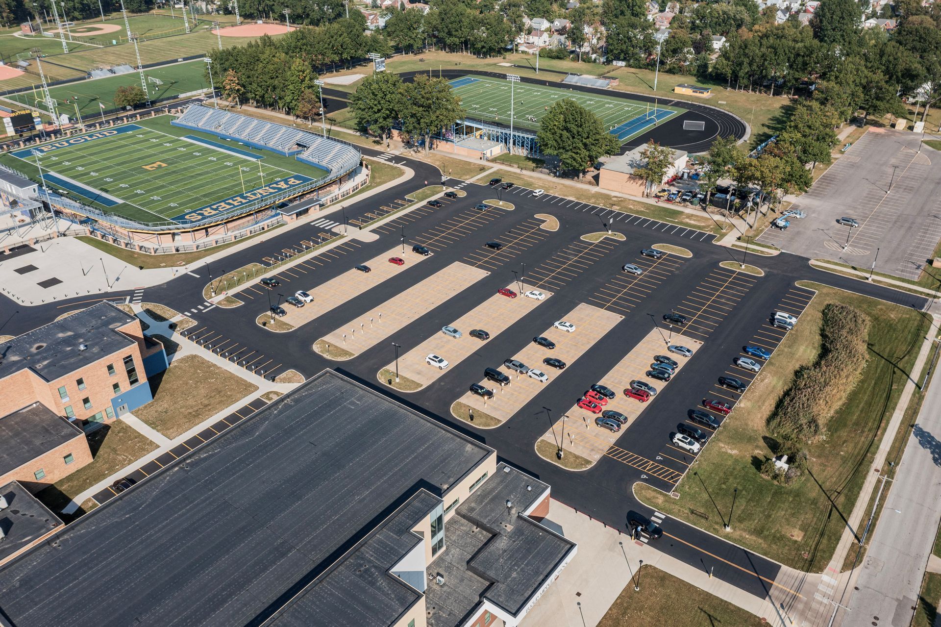 Aerial view of a school with a football field, track, parking lot with cars, and buildings.
