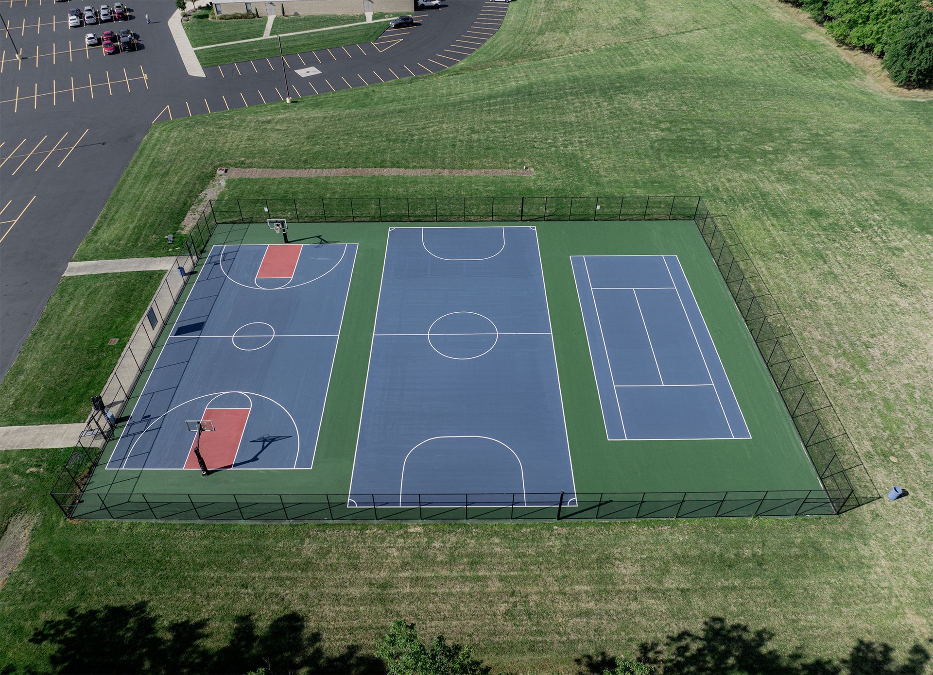 Aerial view of a multi-sport court: basketball court, tennis/pickleball courts, surrounded by a fence and grassy area.