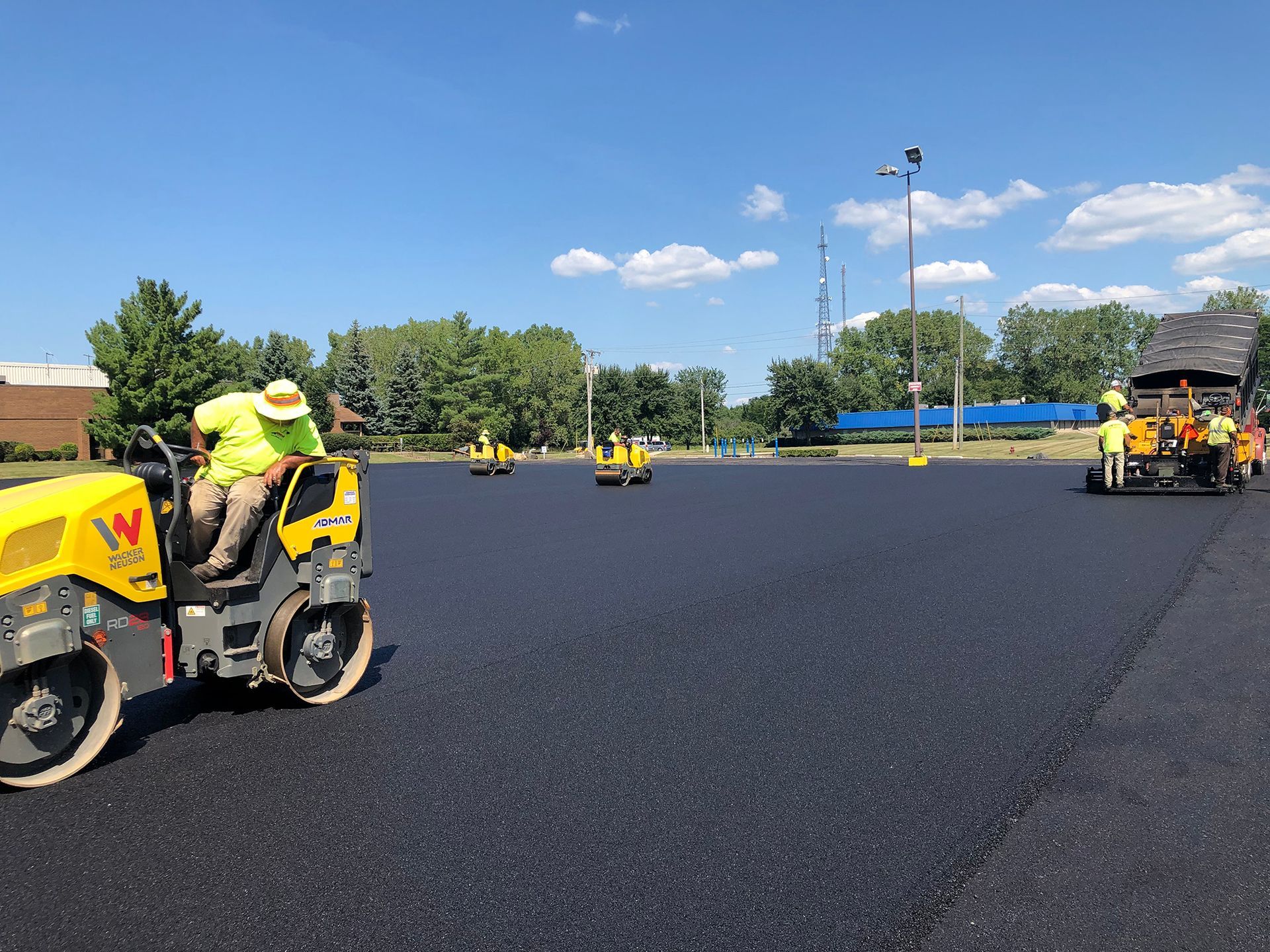 Workers paving asphalt parking lot with machinery on a sunny day.