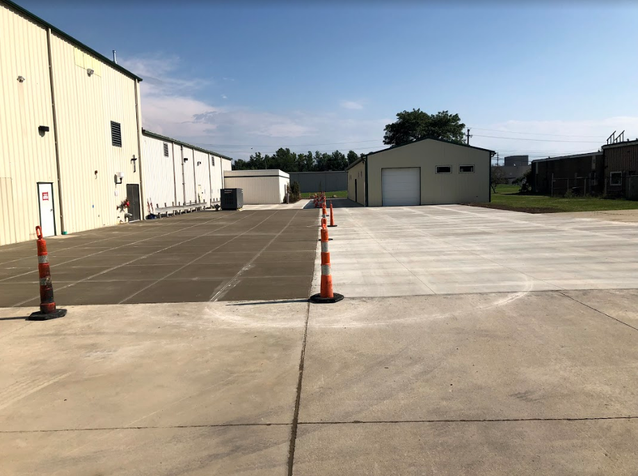 Exterior view of a concrete lot with commercial buildings and orange traffic cones. Blue sky overhead.