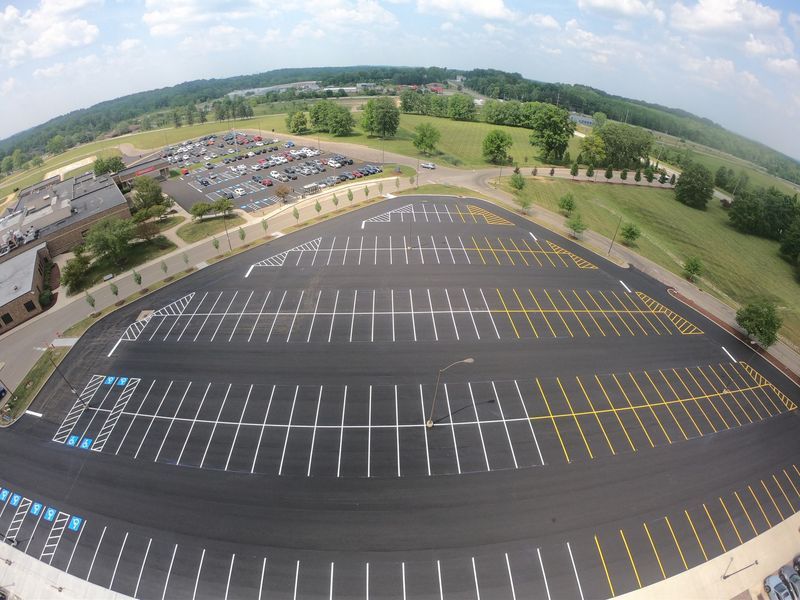 Aerial view of empty parking lot with marked spaces; building and green landscape in background.