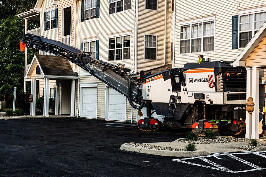 Asphalt milling machine removing pavement near an apartment building with an awning and white doors.