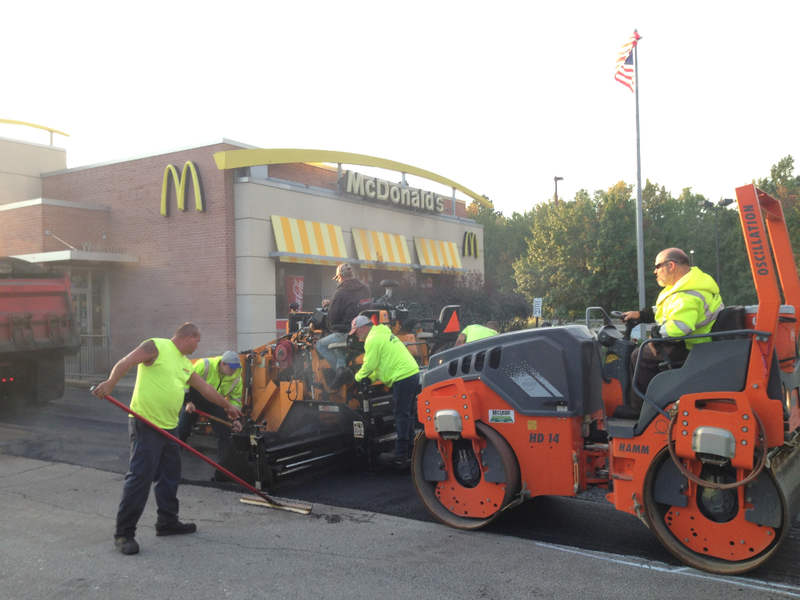 Road crew paving asphalt near a McDonald's restaurant. Workers in neon vests operating machinery under the American flag.