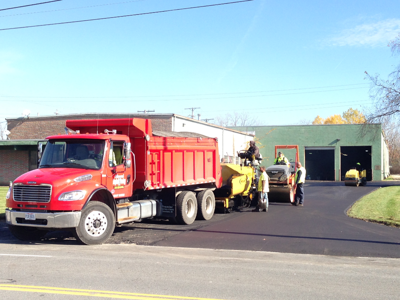 Red dump truck unloading asphalt for a paving crew at a building with green garage doors.
