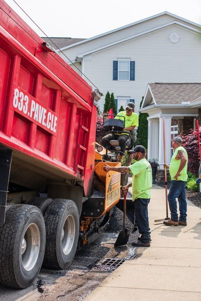Asphalt paving crew, dumping material from red truck into paving machine on residential street.