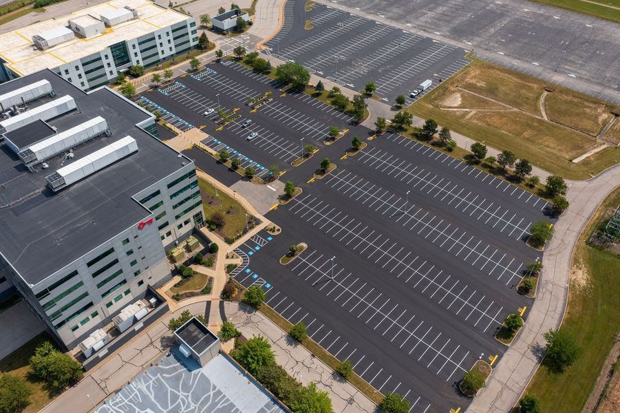 Aerial view of large asphalt parking lots surrounding a modern building. Striped parking spaces are mostly empty.