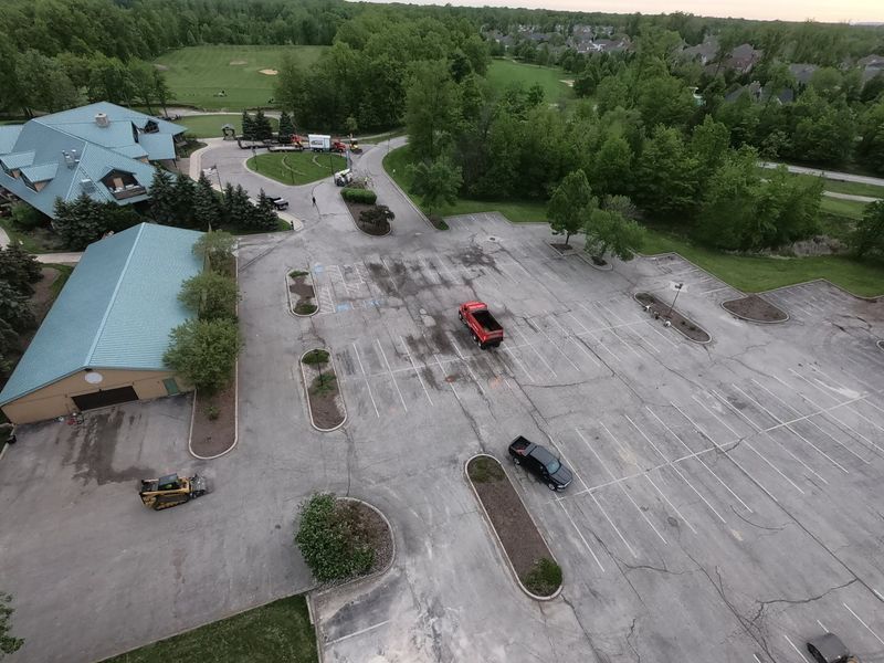 Aerial view of a mostly empty parking lot with a red car and a black car parked. Buildings and trees surround the lot.