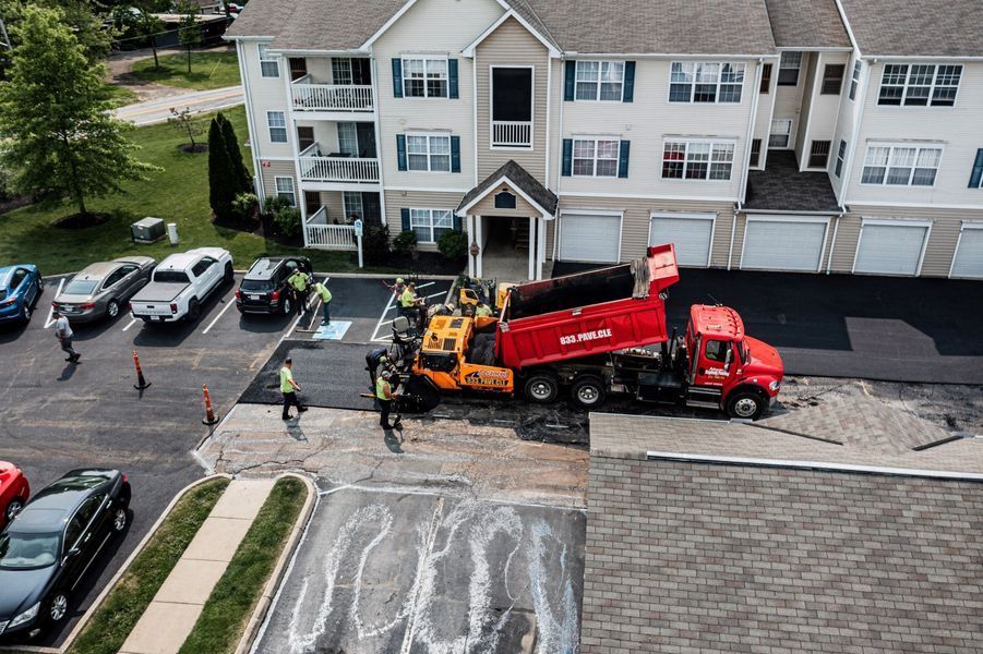 Asphalt paving in a parking lot next to an apartment building; red truck dumping asphalt into a yellow paving machine.