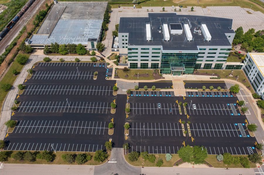 Aerial view of a large office building with expansive parking lots, some trees, and a train track.