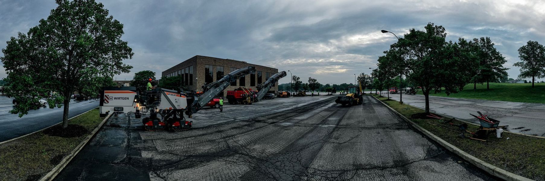 Construction site with building, asphalt work, heavy machinery, cloudy sky, trees.