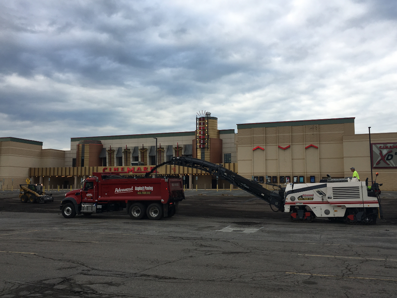 Asphalt milling machine loading truck in front of a theater under cloudy sky.
