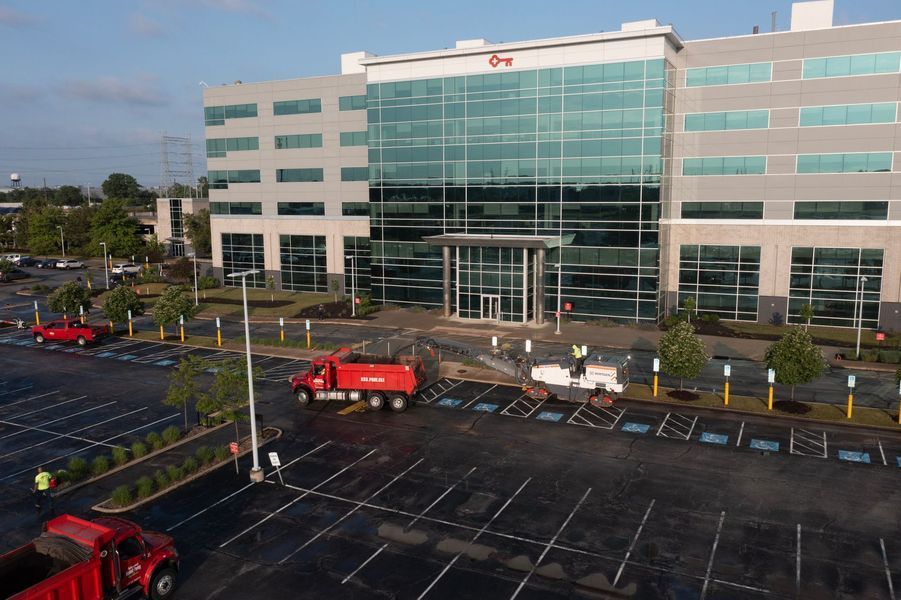 A modern building with glass front, workers near entrance, red dump trucks, and empty parking lot.