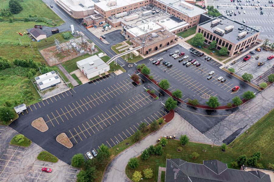 Aerial view of an asphalt parking lot with cars, leading to a beige brick building and surrounding green space.