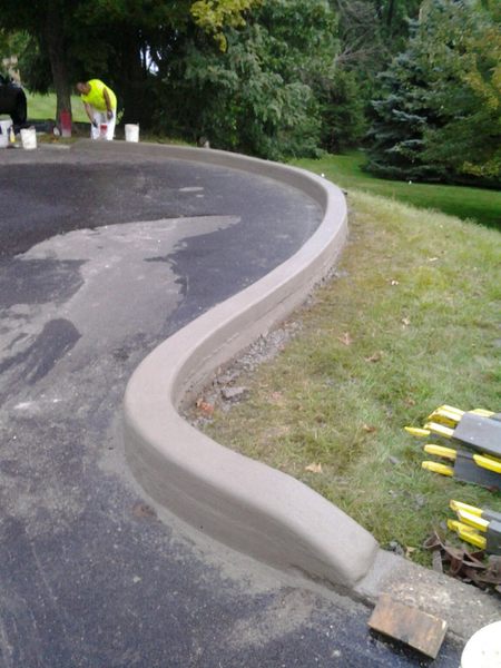 Newly poured curved concrete curb next to asphalt driveway and green grass. A person in a yellow vest is working nearby.