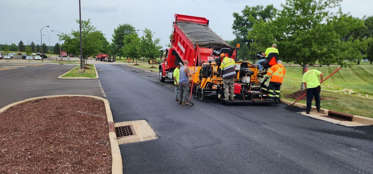 Asphalt paving in progress: truck dumping asphalt into a paver, several workers on site.