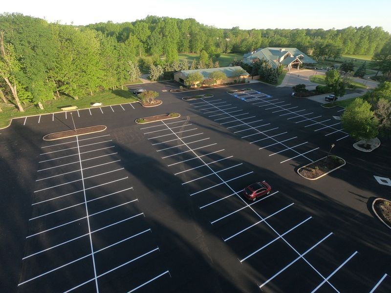 Aerial view of a nearly empty parking lot with white lines, a building in the background, and surrounding trees.