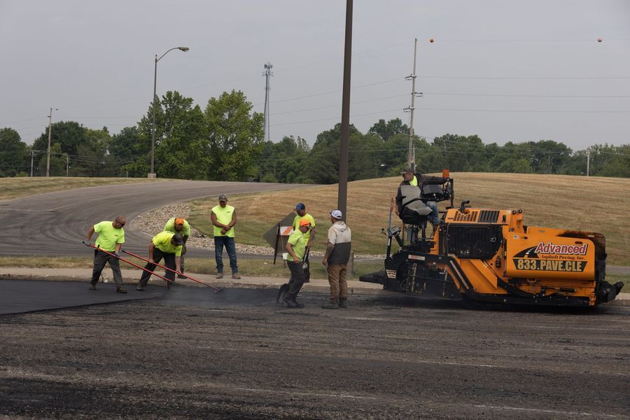 Road workers paving asphalt with a machine; several workers wearing safety vests use tools.