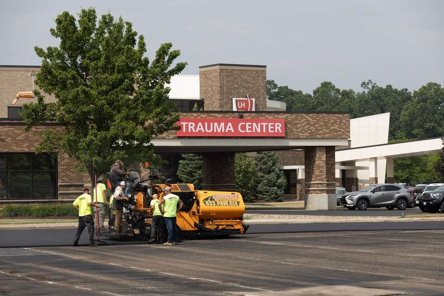 Asphalt paving at a trauma center entrance; workers in vests operate machinery near building with 