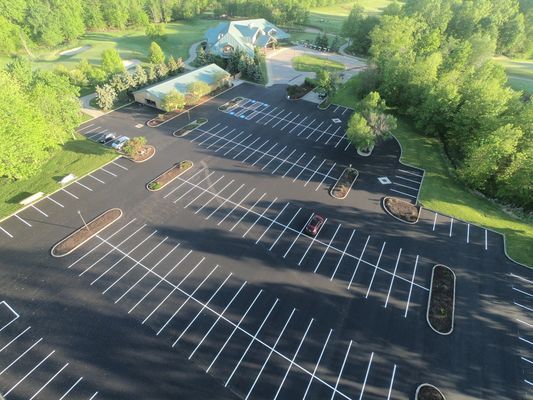 Aerial view of a mostly empty asphalt parking lot with white parking space lines. Trees and a building are in the background.