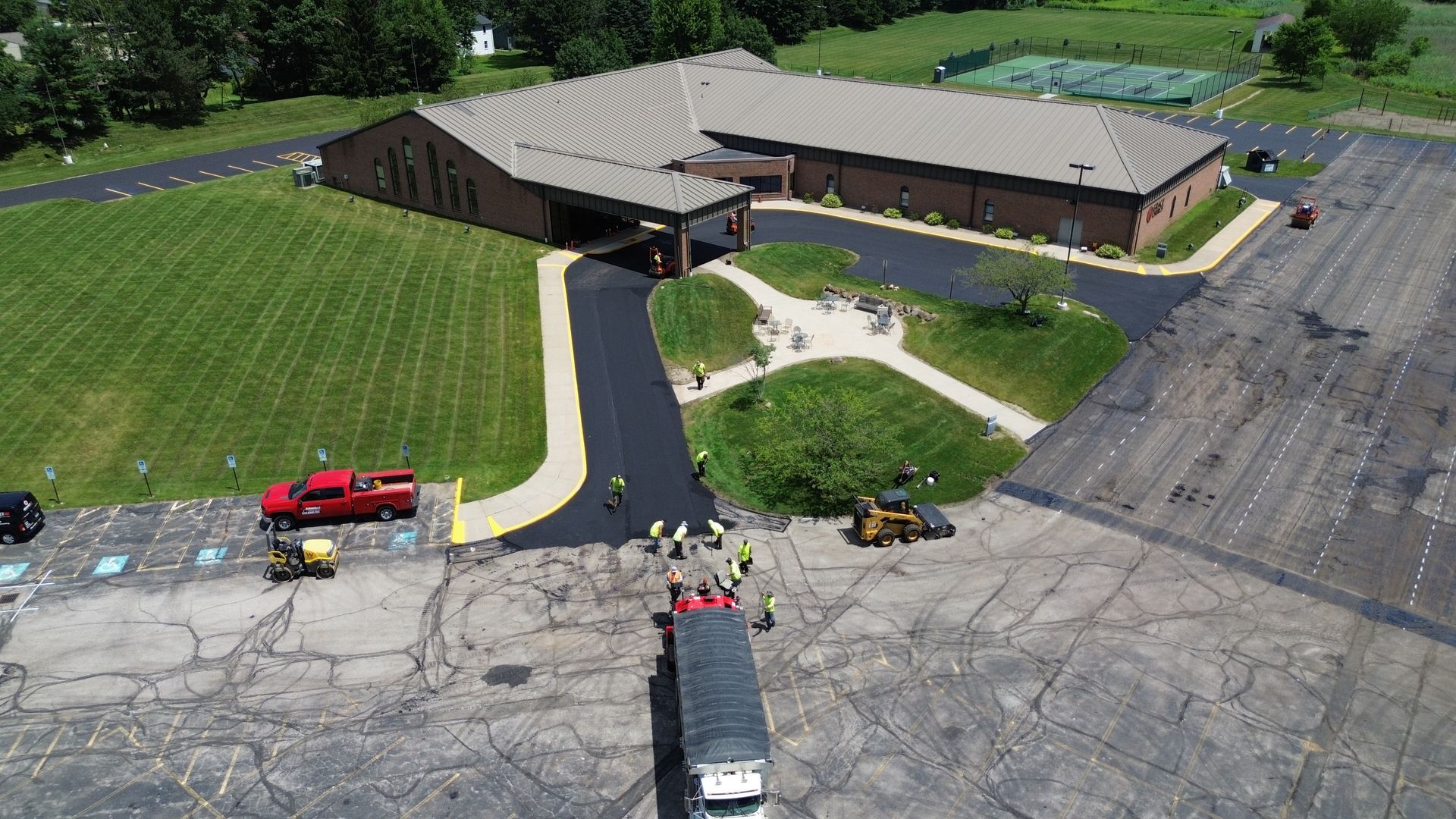 Paving crew working on asphalt in front of a brick building. Red truck, heavy machinery, and workers visible.