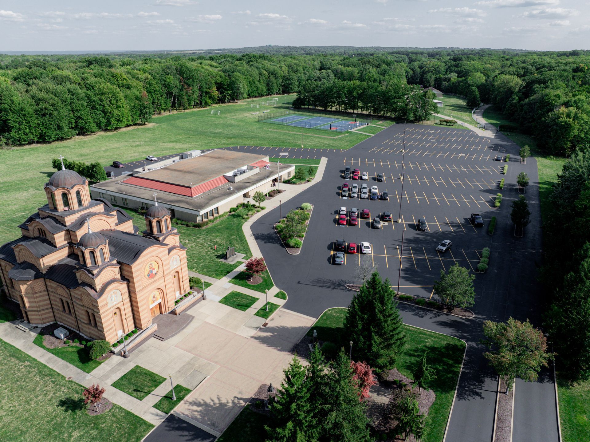 Aerial view of a church and community center with a large parking lot, surrounded by trees and a sports court.