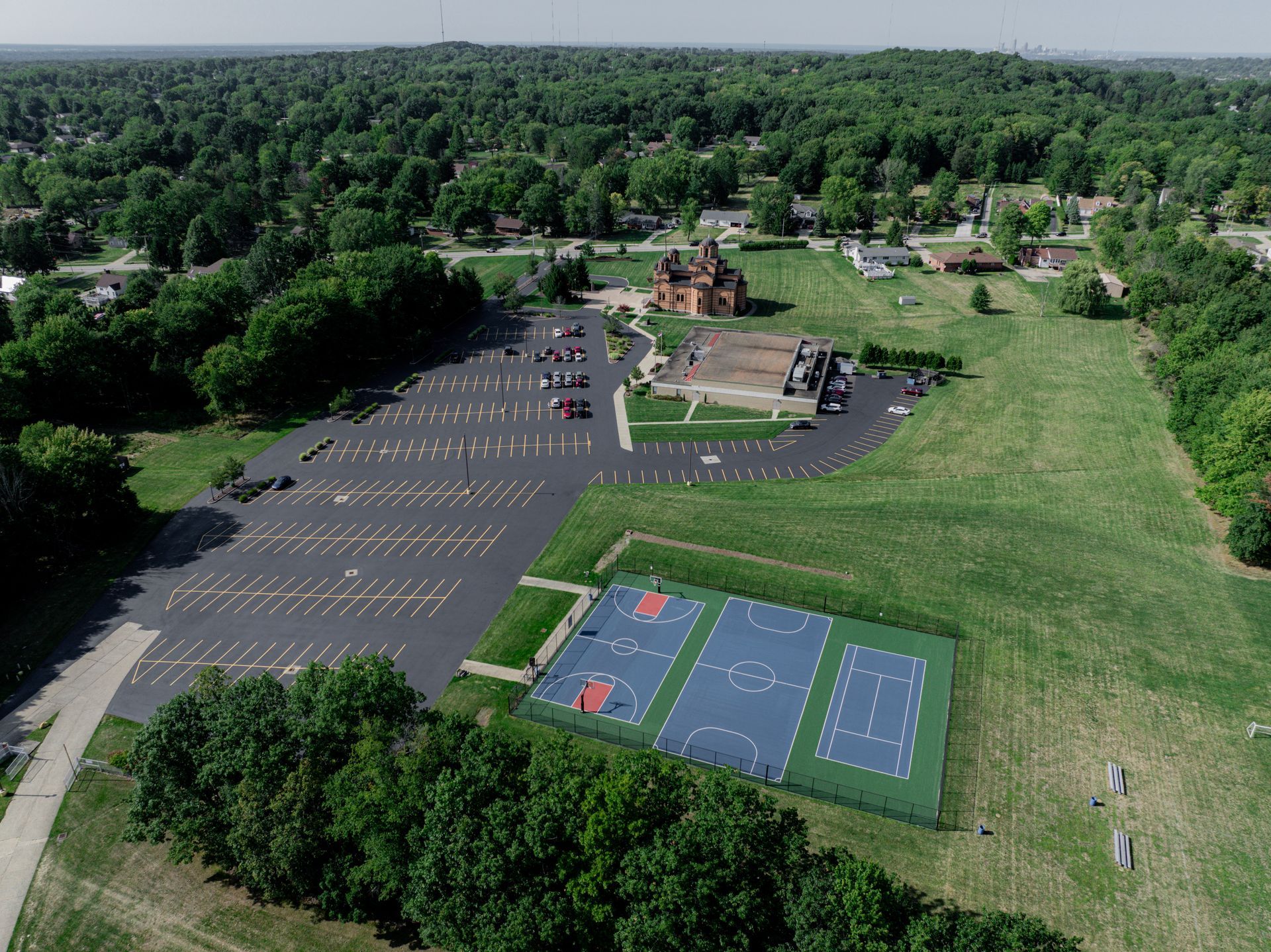 Aerial view of a church and parking lot with two basketball courts and surrounding green space, trees.