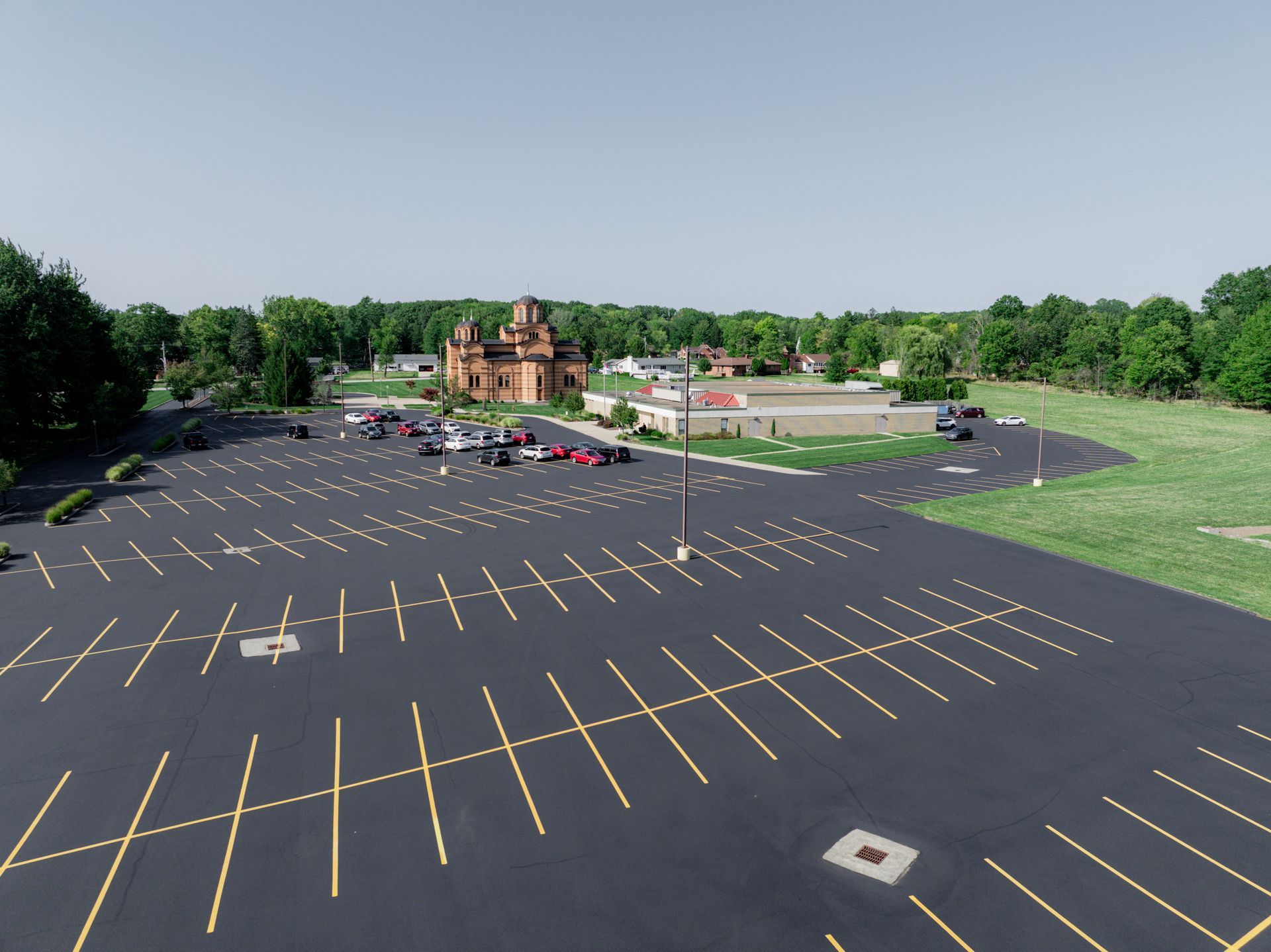 Large empty parking lot with several cars parked in the distance, and a large brick building under a bright blue sky.