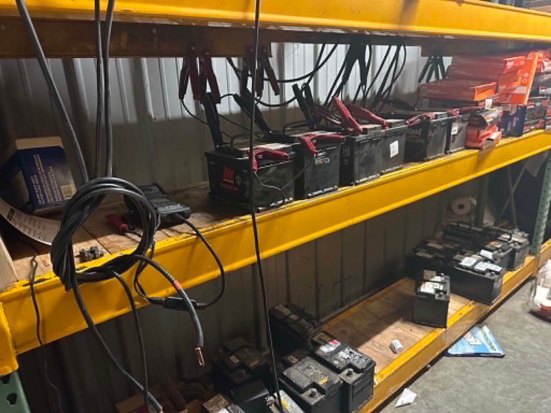Shelf of batteries, cables, and tools in a workshop, with black and red batteries lined up on yellow metal shelves.