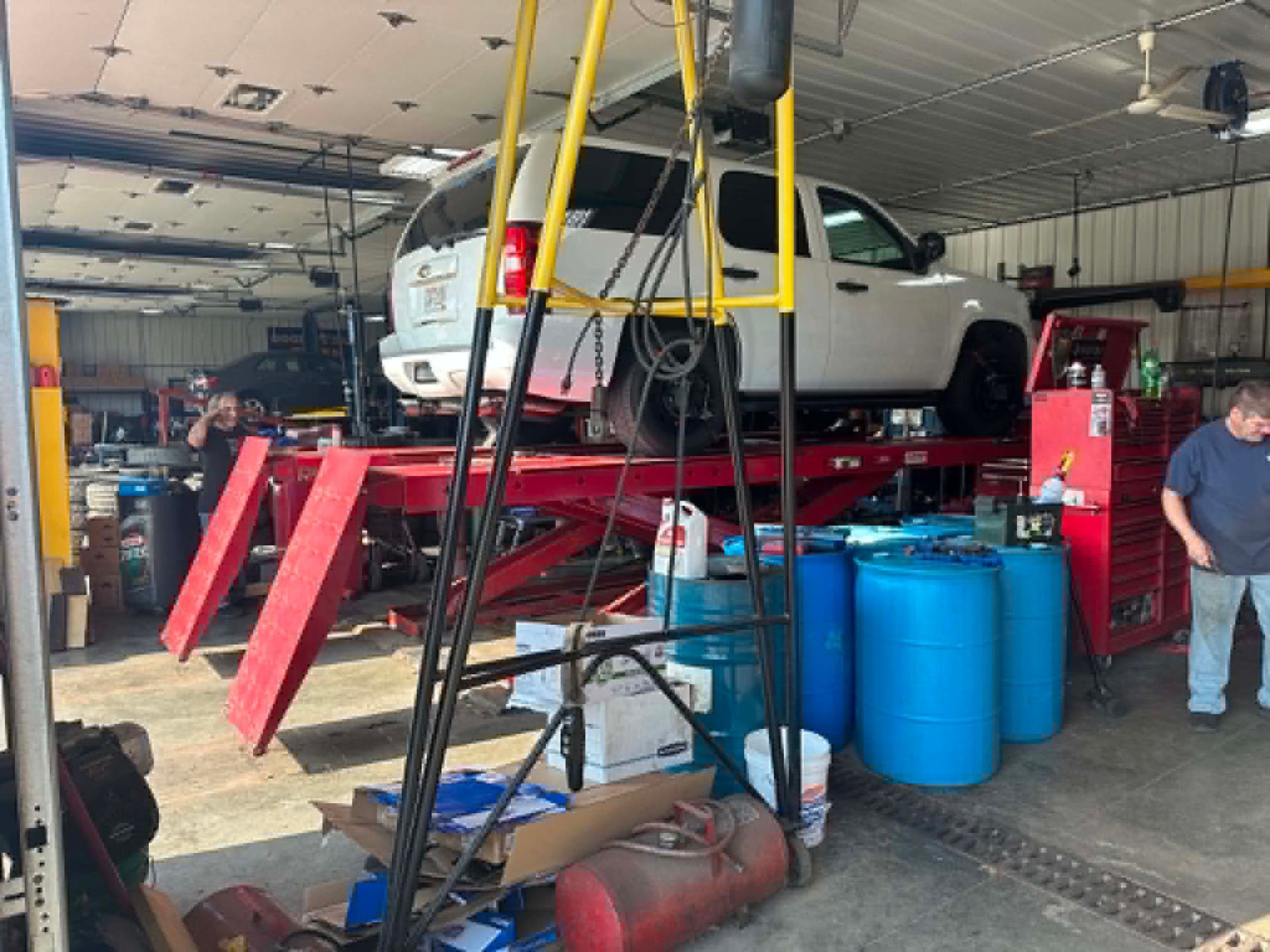 White SUV on a red auto lift in a mechanic’s garage, with blue barrels and tools nearby