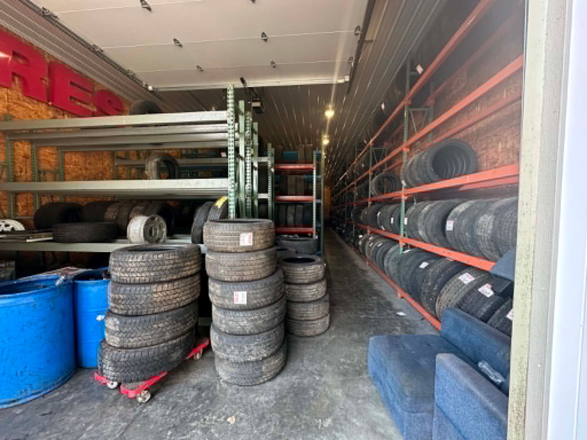 Tire shop storage area with stacks of tires, blue barrels, and shelving in a dim garage.