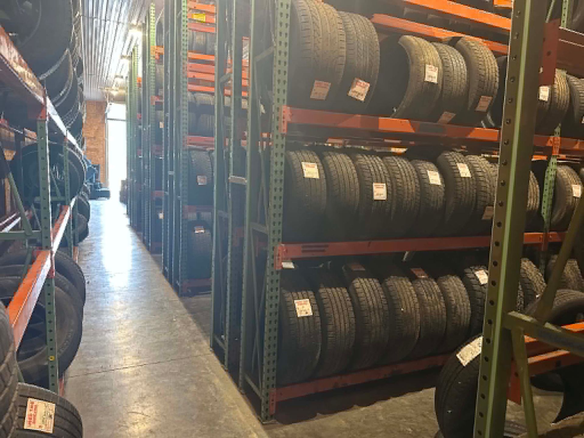 Warehouse aisle lined with stacked tires on metal racks, lit by a bright doorway at the far end