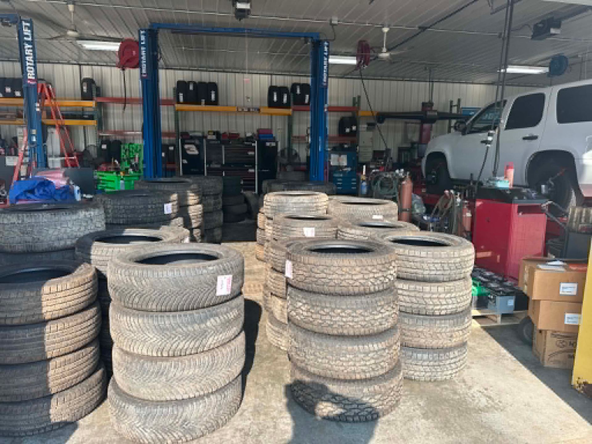 Stacks of used tires in a cluttered auto shop garage with shelves, tools, and a white vehicle in the background