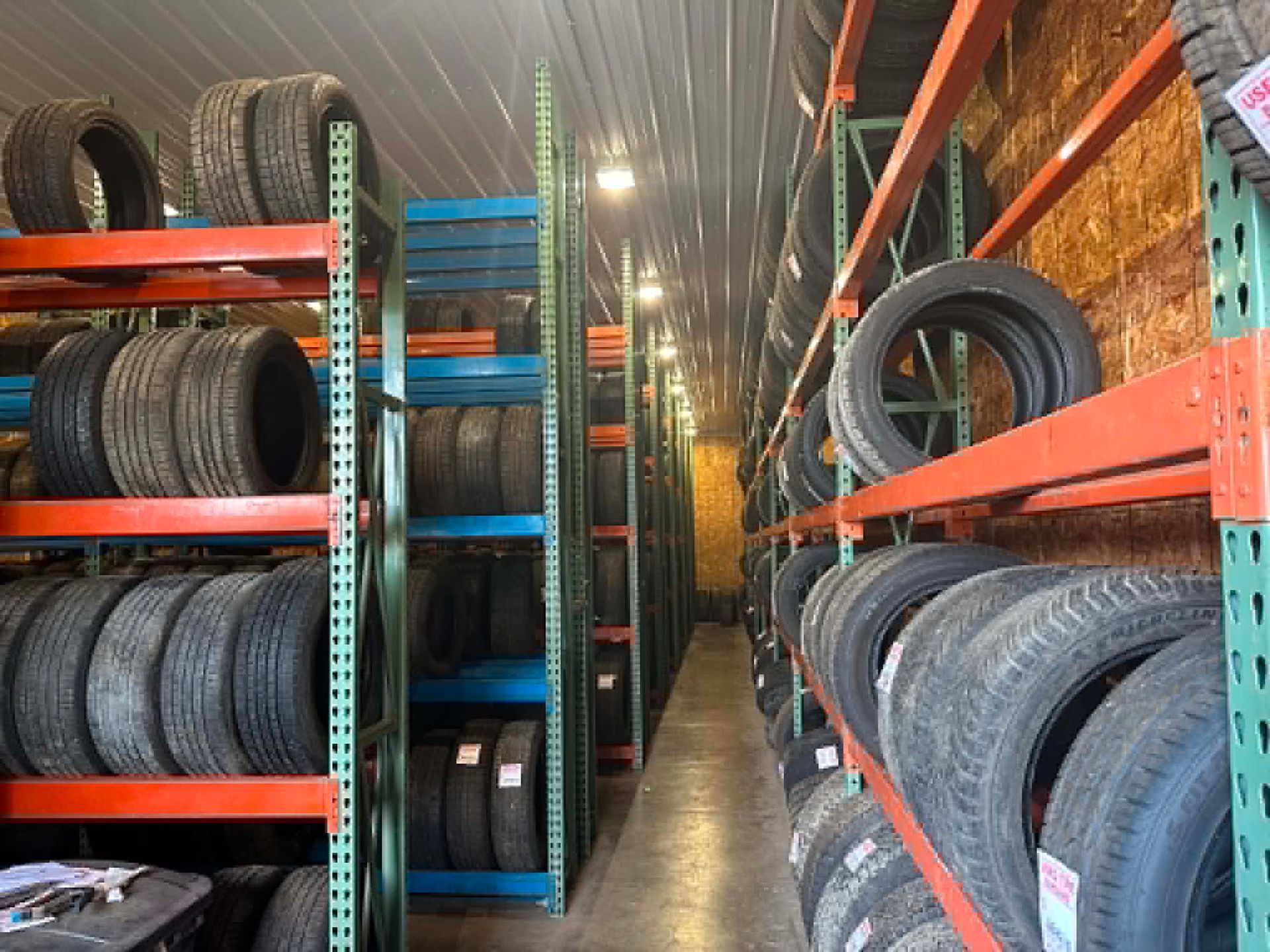 A narrow tire shop aisle with rows of stacked tires on orange and blue metal shelves