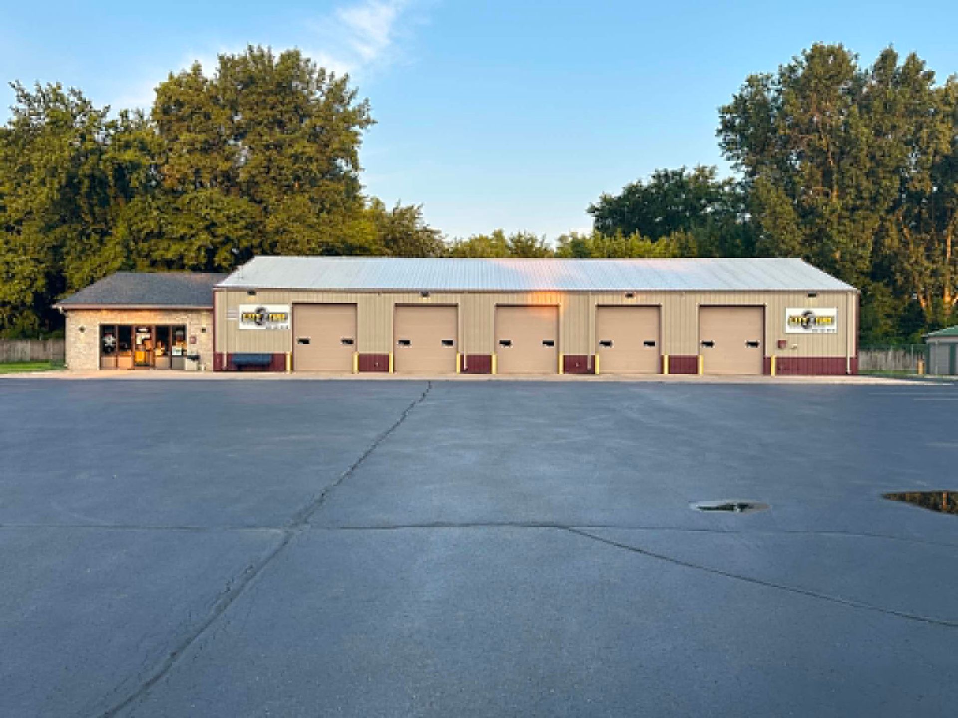 Single-story tan garage building with several doors, beside a large empty paved lot at sunset