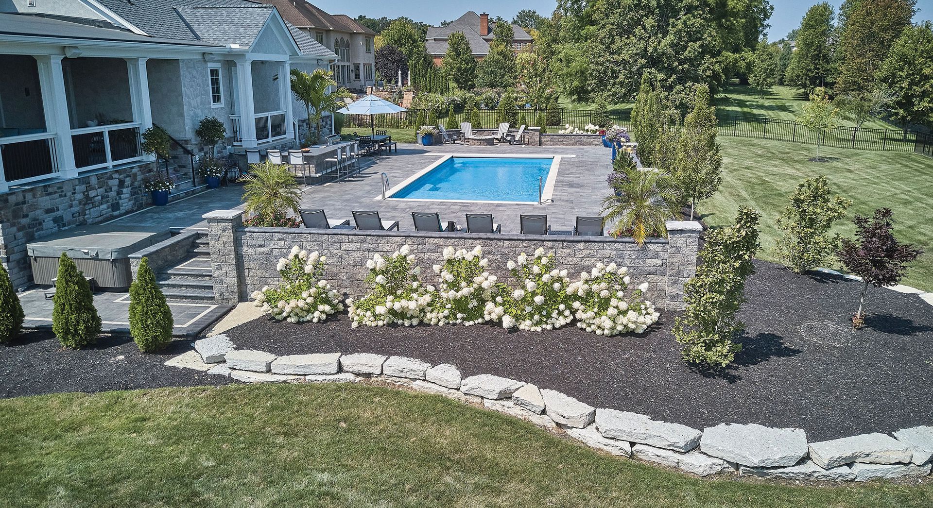 A backyard features a patio with a rectangular swimming pool, lounge chairs, a stone retaining wall, and landscaped beds.