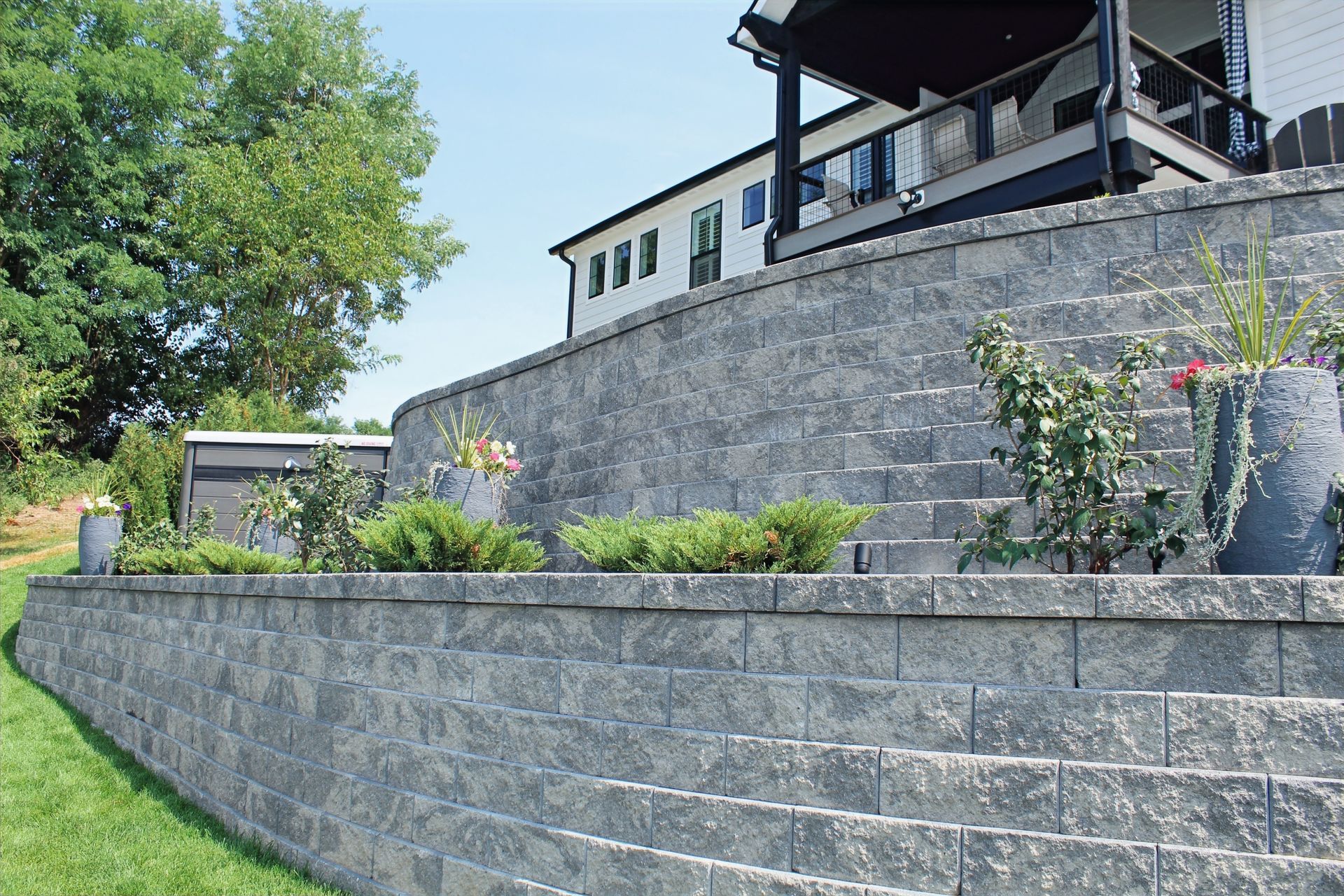 A tall, curved, grey stone retaining wall tiered with green shrubs beneath a modern white house with a dark deck.