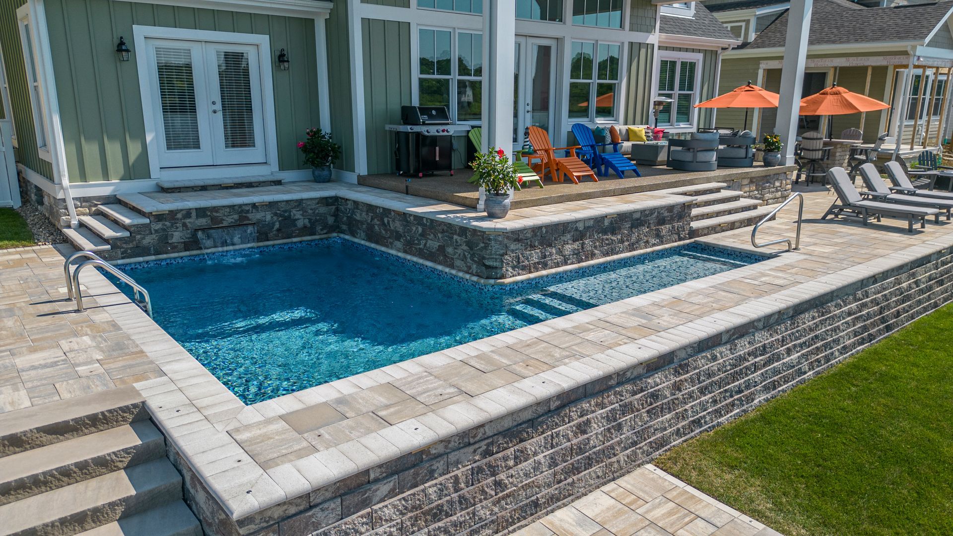 A stone-walled rectangular swimming pool sits next to a patio with colorful chairs and a grill outside a house.