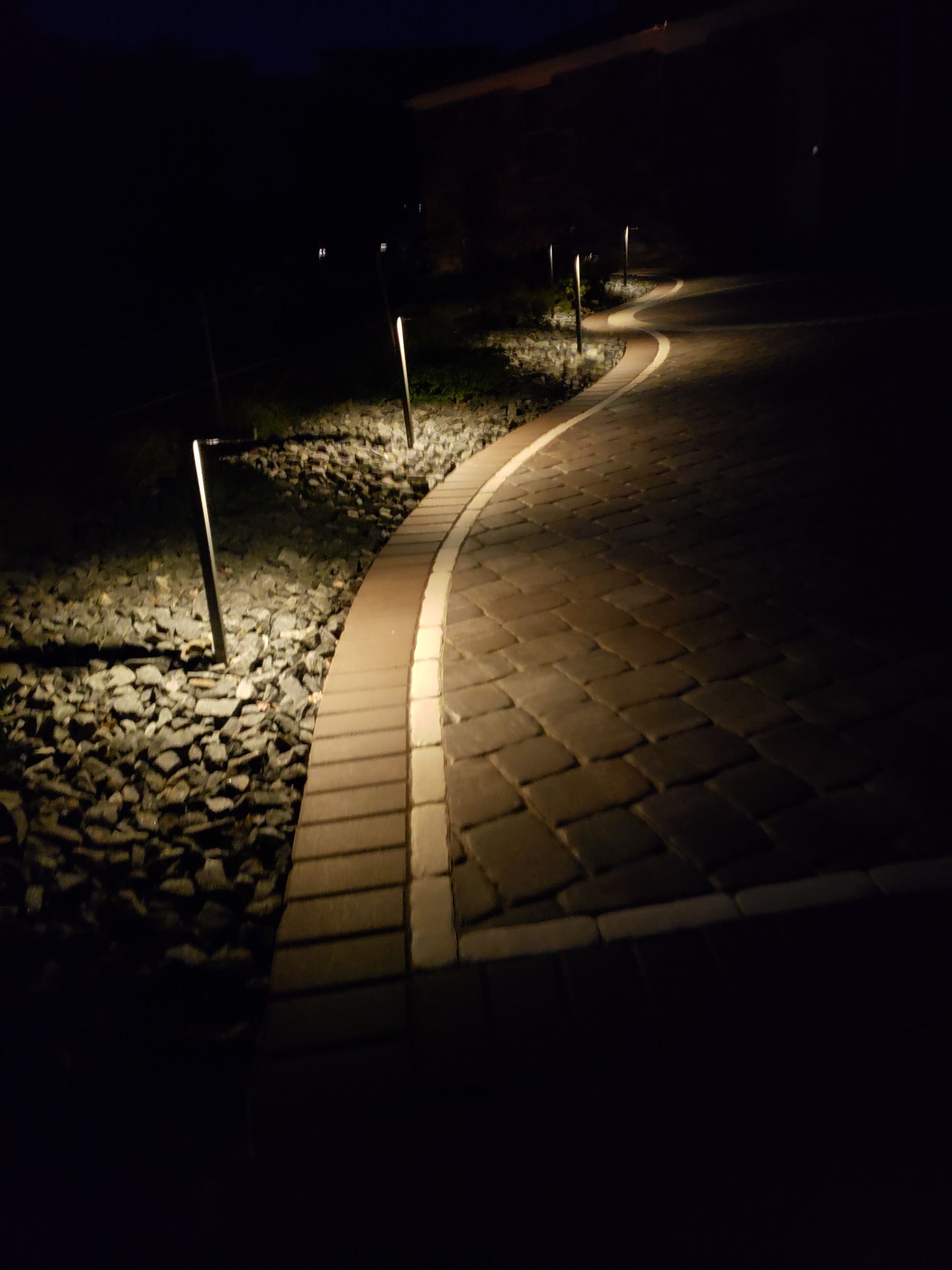 A paved walkway at night, illuminated by a series of small solar stake lights along the edge of a rocky garden bed.