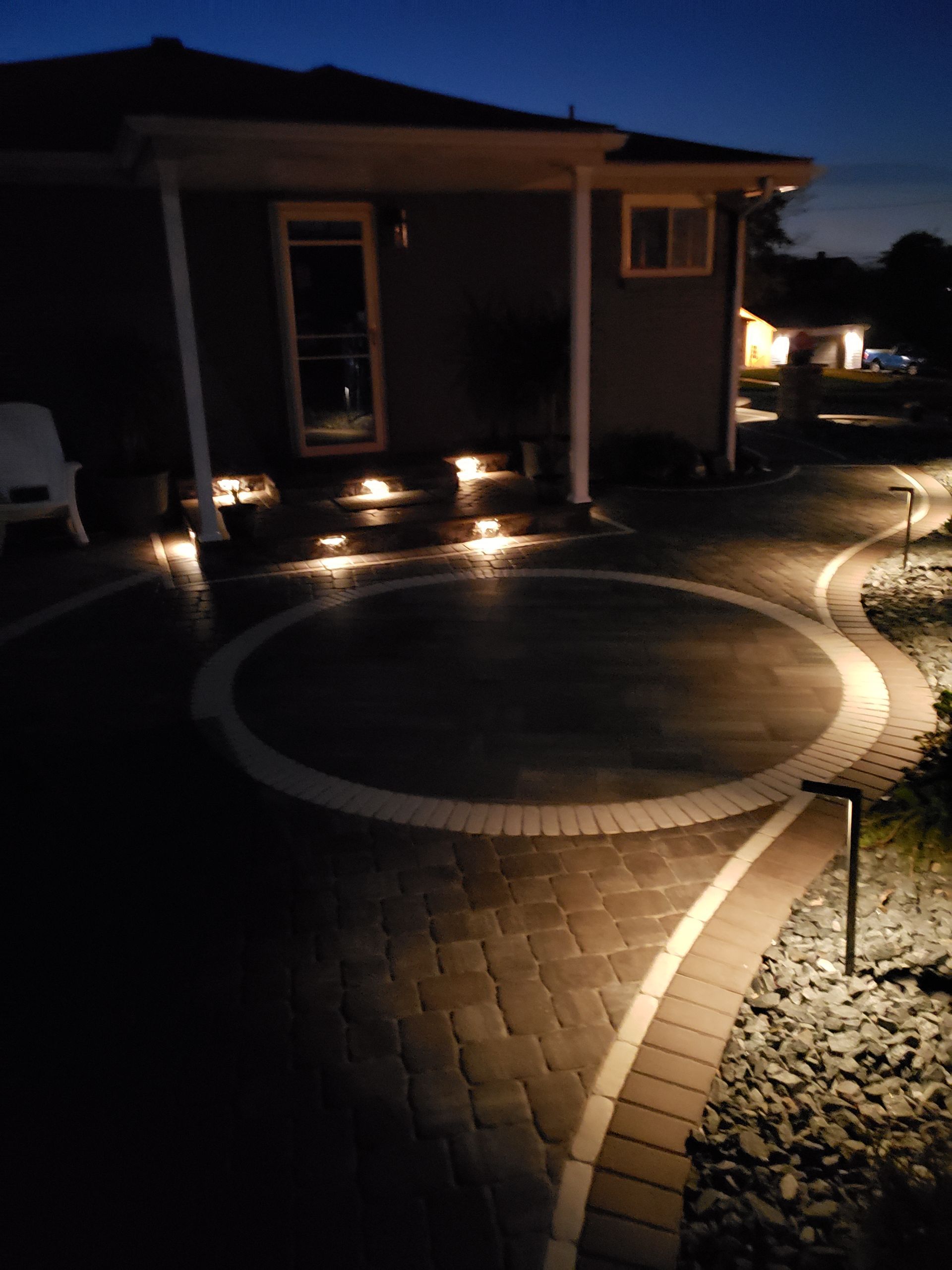 A paved patio with a circular design at night, illuminated by soft step lights and path lighting leading to a house entrance.