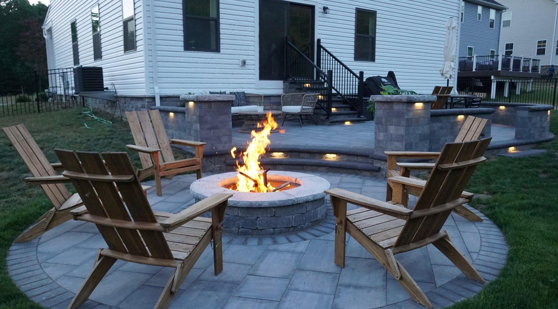 A cozy patio with a glowing fire pit surrounded by four wooden Adirondack chairs, set against a two-story white house.