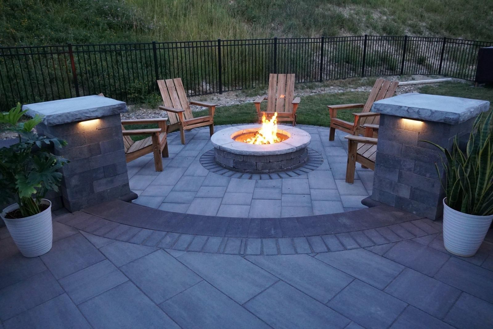 A stone fire pit with a lit fire surrounded by wooden chairs on a paved patio with two stone pillars and potted plants.